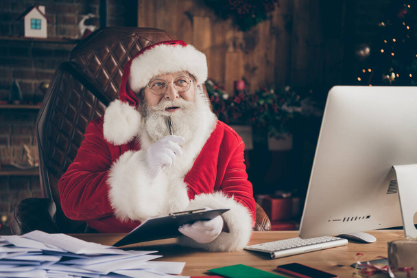 Whait if i. Thoughful minded santa claus sit table prepare x-mas christmas gift order write clipboard think thoughts have crazy newyear ideas wear cap in house indoors christmas tree tinsels - Φωτογραφία, εικόνα