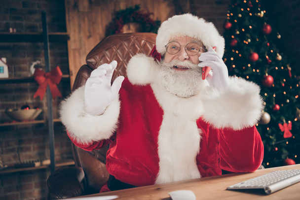 Listen i have x-mas party ideas. Jolly holly white grey hair beard santa claus sit table call smartphone elf tell say christmas news wear cap headwear in house indoors with decoration lights - 写真・画像