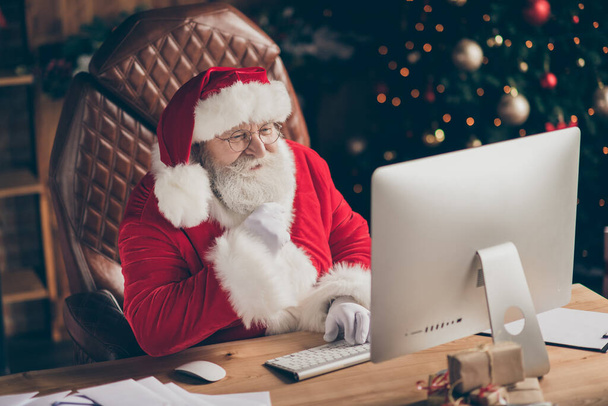 Fairy jolly holly santa claus sit table work computer look screen read wish list letter gift present email touch hand grey beard in house indoors x-mas christmas ornament wear cap headwear - Foto, Imagem
