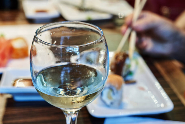 Glass with the remains of white wine on the background of a blurred hand with chopsticks holding sushi - Foto, immagini