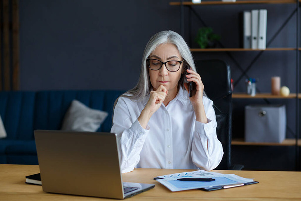 Smiling senior grey-haired beautiful businesswoman talking on the phone in office. Business portrait of attractive mature lady wearing eyeglasses sitting at workplace. Conversation, negotiation - 写真・画像