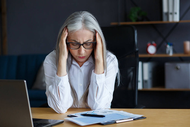 Burnout and overwork concept.Tired businesswoman working overtime in office.Portrait of stressed grey-haired senior woman with head ache.Eye strain after working on laptop.Entrepreneur in bankruptcy - 写真・画像