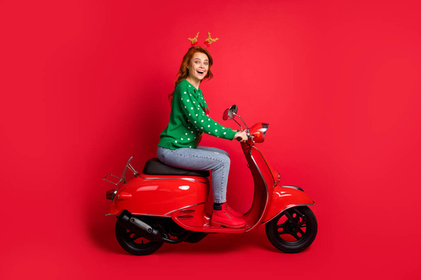 Photo portrait side view of woman riding scooter wearing green jumper deer toy headwear isolated on vivid red colored background - 写真・画像