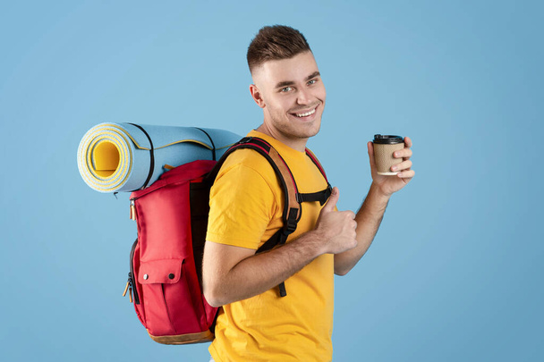Schöner Mann mit Touristenausrüstung, der Kaffee zum Mitnehmen hält und Daumenhoch-Geste über blauem Studiohintergrund zeigt - Foto, Bild
