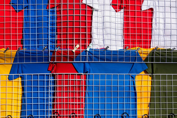 Řádky mnoha barevných triček visí na bílé mřížky sítě oblečení regál na prodej v obchodě s oděvy na venkovním trhu - Fotografie, Obrázek