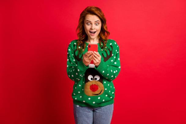 Photo portrait of excited girl with open mouth holding phone in two hands wearing ugly green sweater isolated on vivid red colored background - Photo, Image