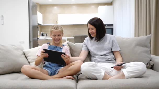 Νεαρή μητέρα και έφηβη κόρη αναπαύονται στον καναπέ στο σπίτι και χρησιμοποιούν τη συσκευή tablet και το smartphone διασκεδάζοντας γελώντας - Πλάνα, βίντεο