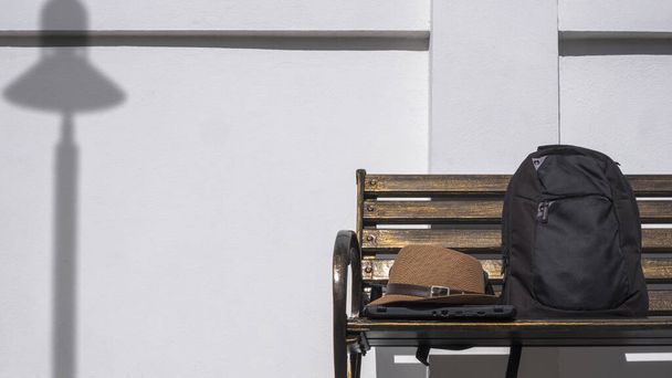 Sombrero fedora vintage con portátil y mochila en banco de parque de metal con sombra de farola en el fondo de la pared de cemento blanco en el área pública - Foto, Imagen