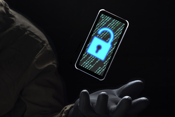 Gros plan illuminer l'icône de déverrouillage avec le code binaire vert à l'écran du smartphone flottant au-dessus de la main du pirate en gant noir sur fond sombre, le piratage et Internet concept de cybercriminalité - Photo, image