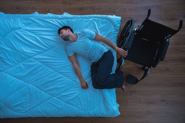 L'uomo sdraiato sul letto vicino a una sedia a rotelle. vista dall'alto, sera notte - Foto, immagini
