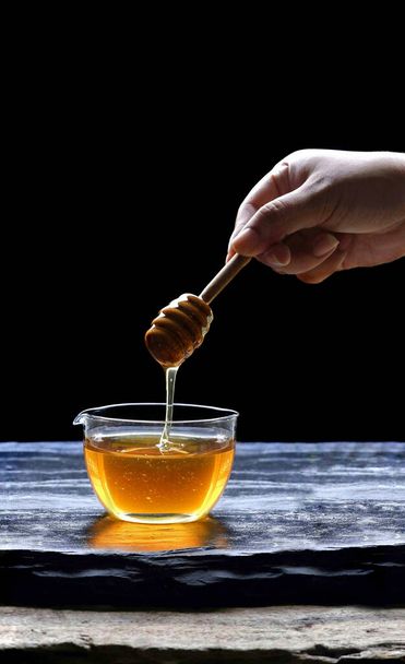 Focus on woman hand holding wooden honey dipper stick and dipping from glass cup on black slate with wooden table in black background,close up and vertical frame - Foto, imagen