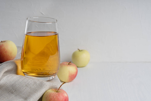 Organic vegetarian apple juice in drinking glass standing on textile towel on white wooden background surrounded by raw ripe fruits showing healthy lifestyle and vegan eating. Image with copy space - Foto, Imagem