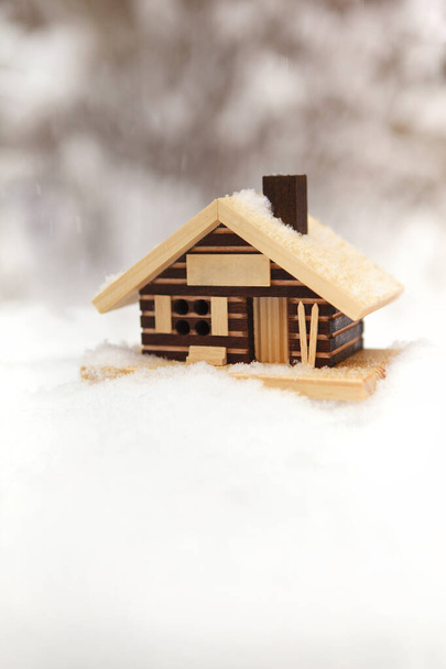 Piccolo modello di casa in legno tra cumuli di neve con nevicate e sfondo foresta invernale offuscata - Foto, immagini