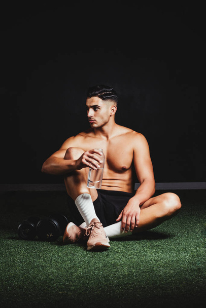 hemdloser muskulöser Mann sitzt auf dem Boden der Turnhalle und trinkt Wasser - Foto, Bild