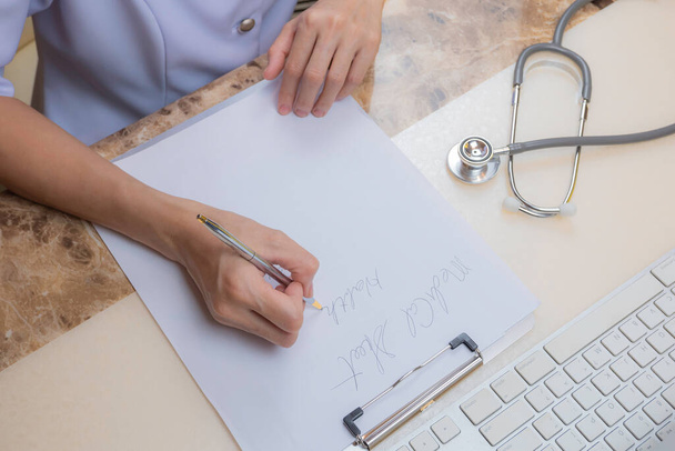 Hohe Blickwinkel der asiatischen Ärztin oder Krankenschwester schreibt Rezept mit Stethoskop und einem Teil der Computertastatur auf den Tisch im Krankenhauszimmer - Foto, Bild