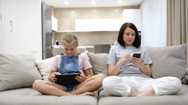 Beyaz tenli genç anne ve kız evdeki kanepede dinleniyor ve tablet cihazını ve akıllı telefonu kullanıyor. Kapalı. - Video, Çekim