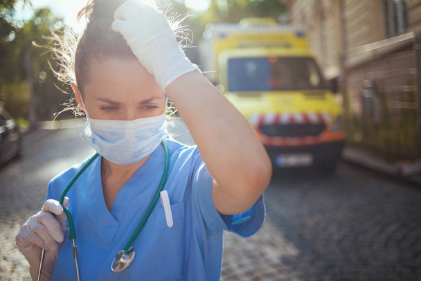 coronavirus pandemic. sad modern medical doctor woman in scrubs with stethoscope and medical mask outdoors near ambulance. - Фото, зображення