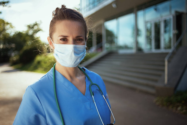 covid-19 pandemic. Portrait of modern physician woman in uniform with stethoscope and medical mask outside near clinic. - Фото, зображення