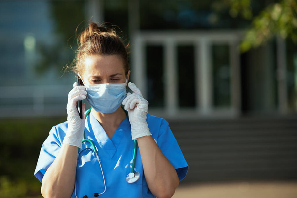 covid-19 pandemic. modern medical doctor woman in scrubs with stethoscope and medical mask speaking on a smartphone outside near hospital. - Фото, зображення