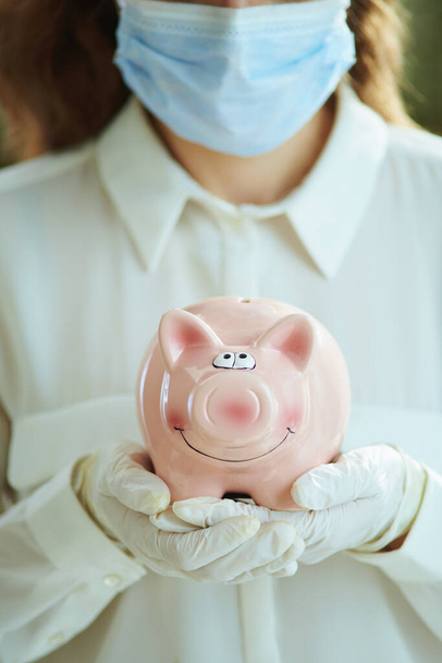 Life during covid-19 pandemic. Closeup on woman in white blouse with medical mask, piggy bank and gloves. - Photo, image