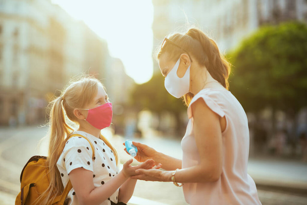 Life during covid-19 pandemic. modern mother and daughter with masks and yellow backpack disinfecting hands with an antibacterial agent and getting ready for school outdoors. - Foto, afbeelding