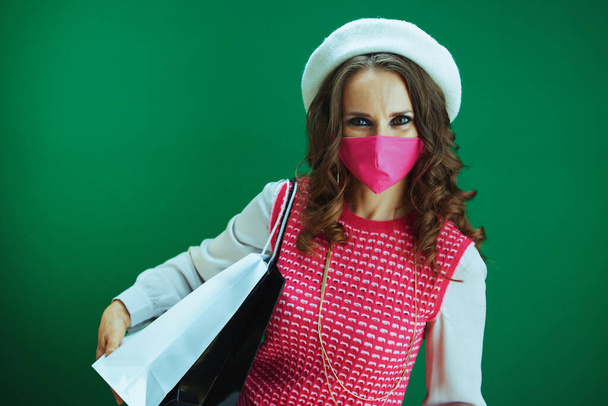 Life during coronavirus pandemic. Portrait of happy modern woman in pink sleeveless shirt with pink medical mask and shopping bags against green background. - Фото, зображення