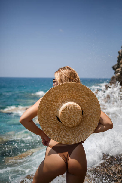 όμορφη νεαρή γυναίκα με καφέ μαγιό και καπέλο περπατά κατά μήκος της παραλίας δίπλα στη θάλασσα - Φωτογραφία, εικόνα