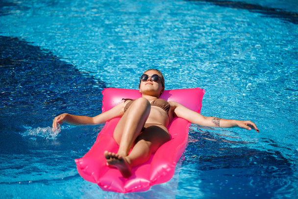 Havuzdaki pembe şişme yatakta mayo giymiş seksi bir kadın yatıyor. Sıcak ve güneşli bir yaz gününde havuz kenarında rahatla. Tatil kavramı - Fotoğraf, Görsel
