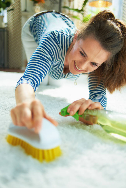smiling modern middle age housewife in striped t-shirt and white pants with spray bottle of eco friendly cleaning supplies and yellow brush spot cleaning white carpet at modern home in sunny day. - Foto, immagini