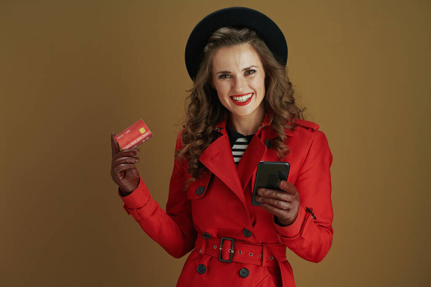 Hello november. smiling modern woman in red coat and black beret with smartphone, credit card and leather gloves on brown background. - Photo, image