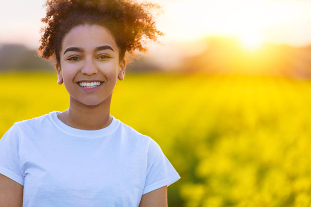 Beautiful smiling mixed race African American girl teenager female young woman with perfect teeth at sunset or sunrise in a field of yellow flowers - Foto, afbeelding