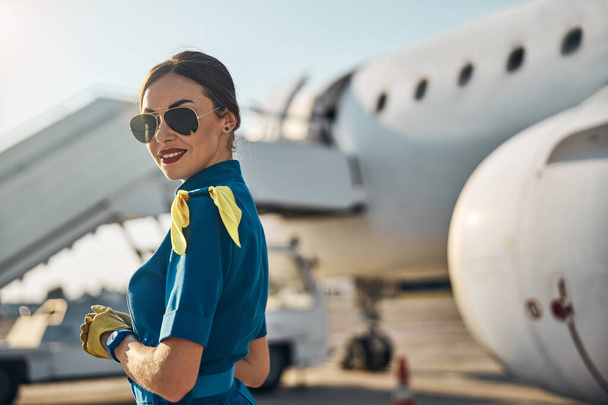 Joyful beautiful stewardess standing near jet outdoors - Foto, imagen