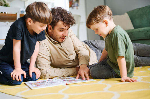 Батько з синами читає книгу, проводячи час разом вдома і лежачи на підлозі
 - Фото, зображення