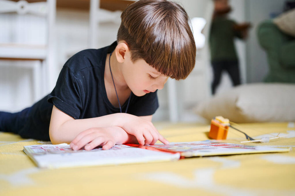 Biały chłopiec z przedszkola leżący w domu na podłodze, czytający książki i oglądający zdjęcia. Koncepcja edukacji domowej - Zdjęcie, obraz