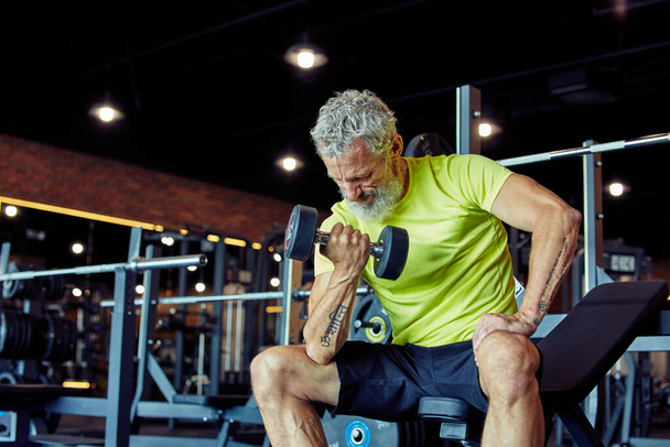 Крепкие руки. Сосредоточенный взрослый мужчина в спортивной одежде, поднимающий гантели и качающий бицепсы во время тренировки в спортзале - Фото, изображение