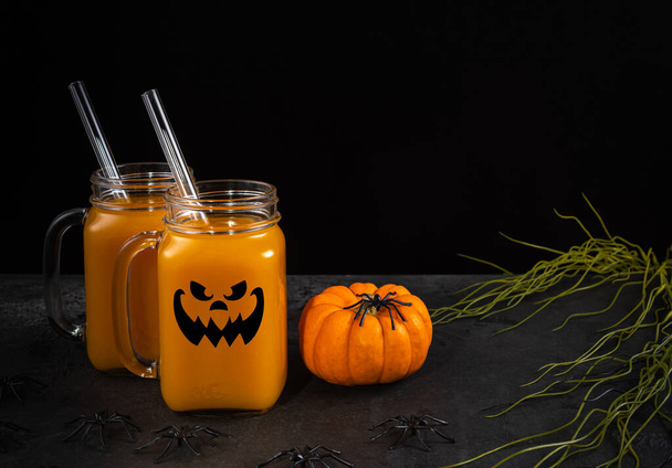 Glass jars with halloween face, pumpkin mocktail and glass straws on black textured table decorated with spiders, spooky plants and pumpkin. Horizontal background with copy space. Selective focus. - Foto, Imagem