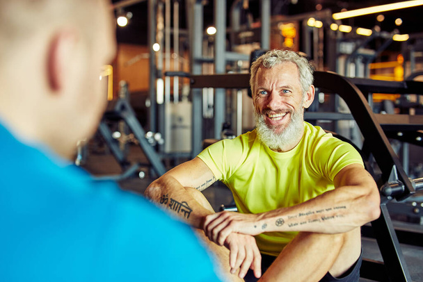 Portret szczęśliwego mężczyzny w średnim wieku omawiającego wyniki treningu z instruktorem fitness lub osobistym trenerem i uśmiechającego się siedząc razem na podłodze na siłowni - Zdjęcie, obraz