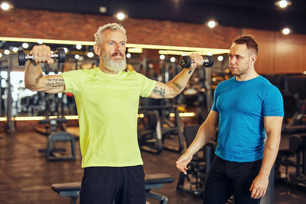 Training body in any age. Focused mature caucasian man in sportswear exercising with dumbbells under supervision of a young professional personal trainer at gym - 写真・画像