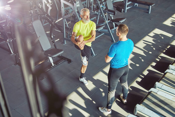 Exercising with personal trainer. Middle aged man in sportswear warming up, stretching his legs and talking with fitness instructor at gym or sport club - Photo, Image