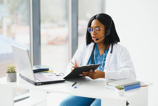 African doctor wear headset consult female black patient make online webcam video call on laptop screen. Telemedicine videoconference remote computer app virtual meeting. Over shoulder videocall view. - Foto, afbeelding