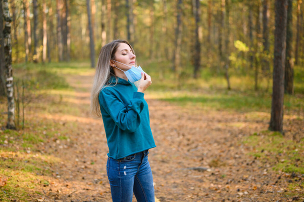 A rapariga está na floresta com uma máscara médica. A menina tira a máscara e respira ar fresco na floresta de outono. - Foto, Imagem