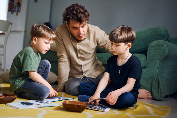 Молодий кавказький батько проводить час вдома з синами і читає книги на підлозі. Щасливий батько грає з дітьми дошкільного віку. Концепція домашньої освіти
 - Фото, зображення
