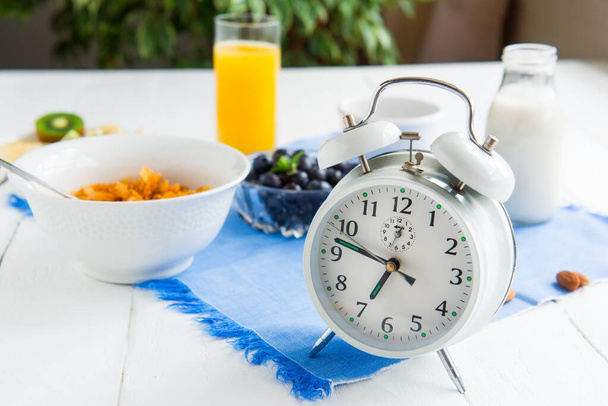 Time for breakfast. Alarm clock with healthy food on the background. Bowl with cereals, nuts and berries, milk, fresh juice on blue napkin and white wooden table. Day diet planning and healthy eating. - Foto, Imagen