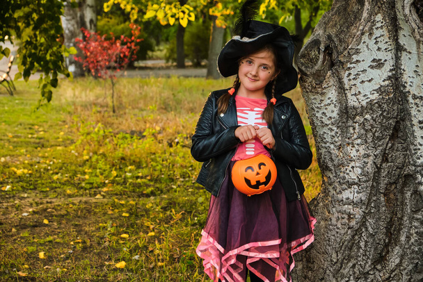秋の公園で魔女の帽子の少女。ハロウィーンのトリックや治療で楽しんでいる子供。子供のトリックや治療。ジャック・オランタンの幼児。秋の森の中でお菓子のバケツを持つ子供. - 写真・画像
