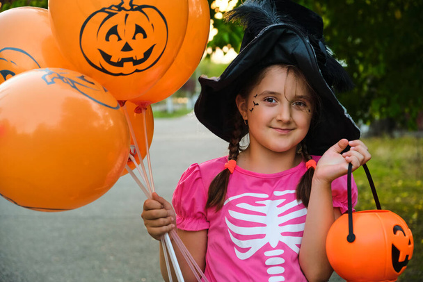 Zabawna dziewczynka Halloween z makijażem na twarzy w strojach karnawałowych na zewnątrz. Portret uśmiechnięta dziewczyna w kapeluszu czarownicy z pomarańczowymi balonami. - Zdjęcie, obraz