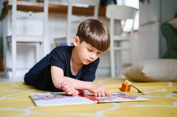 Anaokulu beyaz çocuğu evde yerde uzanıyor, kitap okuyor ve resim seyrediyor. Ev eğitimi kavramı - Fotoğraf, Görsel