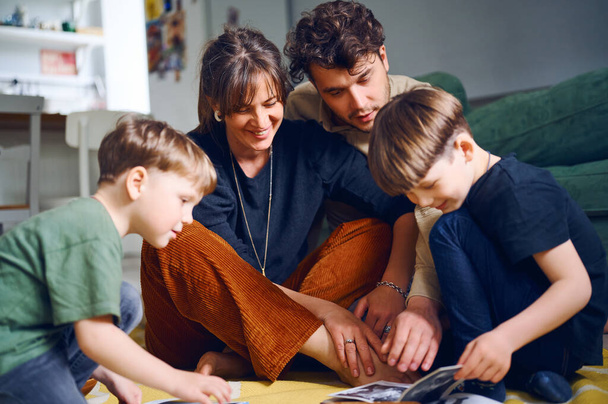 Νεαροί καυκάσιοι γονείς περνούν χρόνο στο σπίτι με γιους και διαβάζουν βιβλία στο πάτωμα. Ευτυχισμένη οικογένεια παίζει με παιδιά preshool. Έννοια της εκπαίδευσης - Φωτογραφία, εικόνα