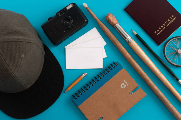Beyaz kartvizitli siyah kamera, gri şapkalı davul çubuğu, bisiklet tekerli fırça, not defteri ve mavi arka planda bir pasaport. - Fotoğraf, Görsel