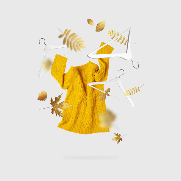 Geel oranje vliegende vrouwen gebreide trui wit houten hangers gouden herfst bladeren op grijze achtergrond. Herfst verkoop Creatieve kleding concept trendy herfst winter gezellige trui trui. Vrouwelijke mode - Foto, afbeelding