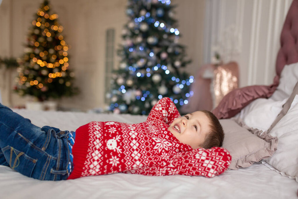 Happy little boy in red knitted sweater, dream on about presents laying in the bed. Christmas tree background. Enjoy winter holidays - Foto, Bild